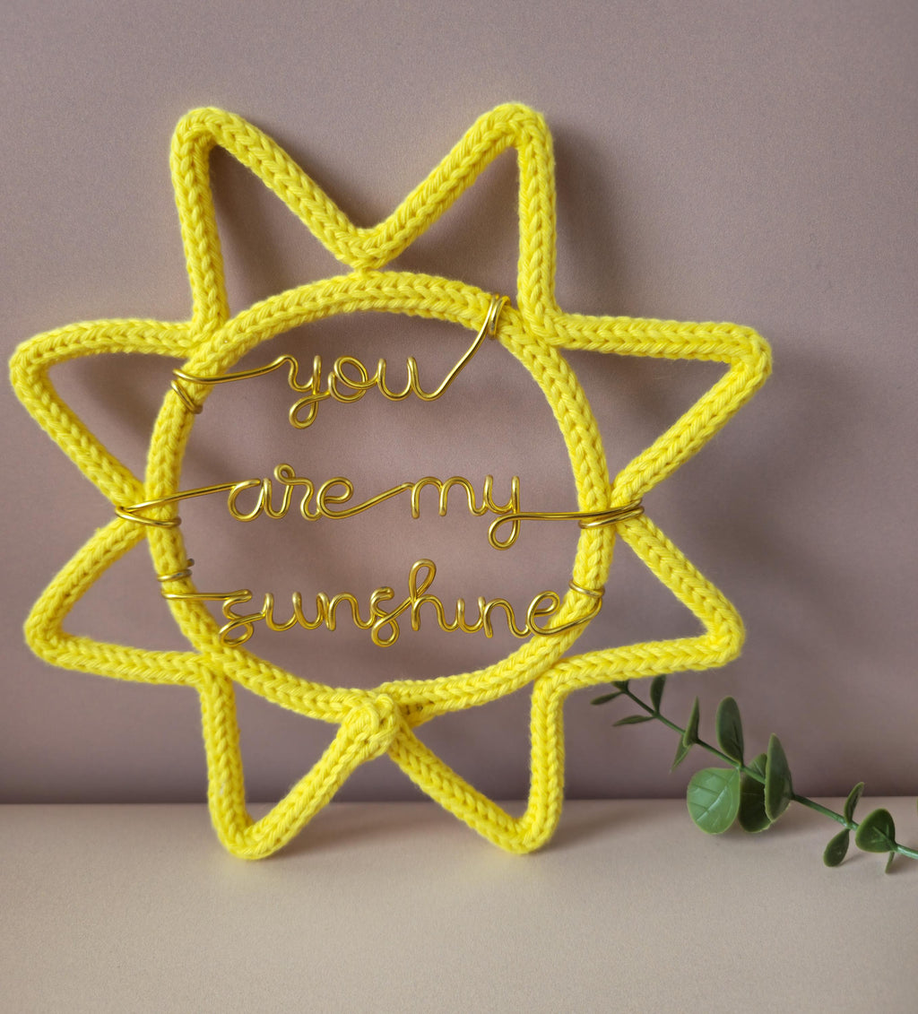 Yellow knitted sun-shaped decoration with 'you are my sunshine' text on a neutral background