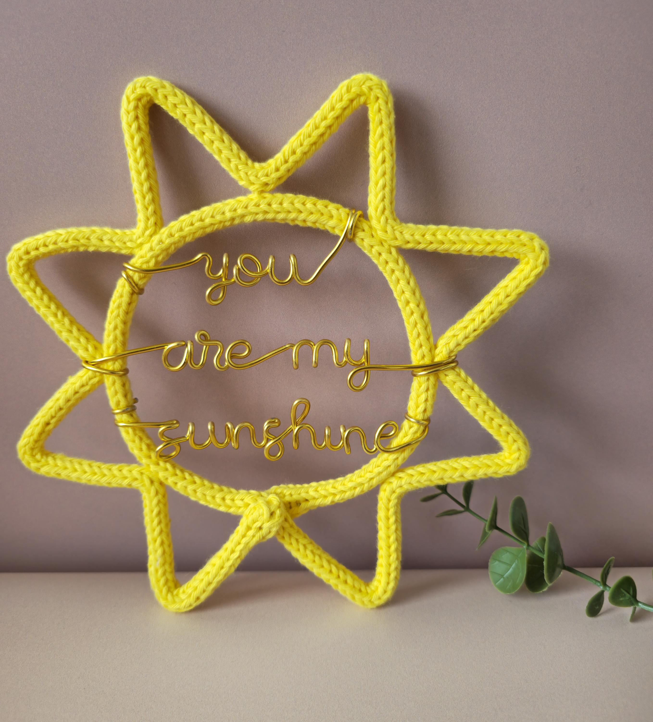 Yellow knitted sun-shaped decoration with 'you are my sunshine' text on a neutral background