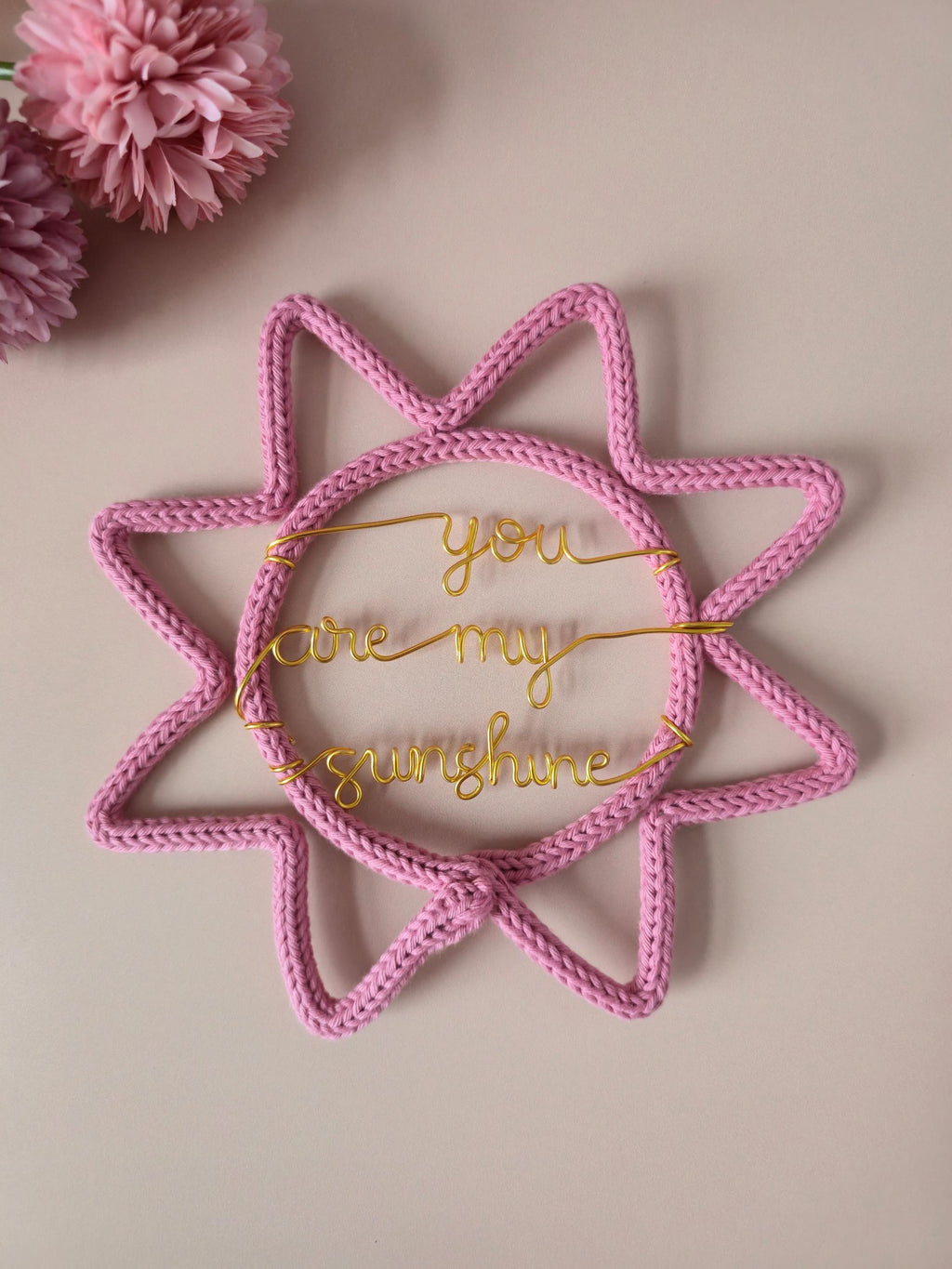 Pink knitted sun-shaped decoration with 'you are my sunshine' text on a beige background