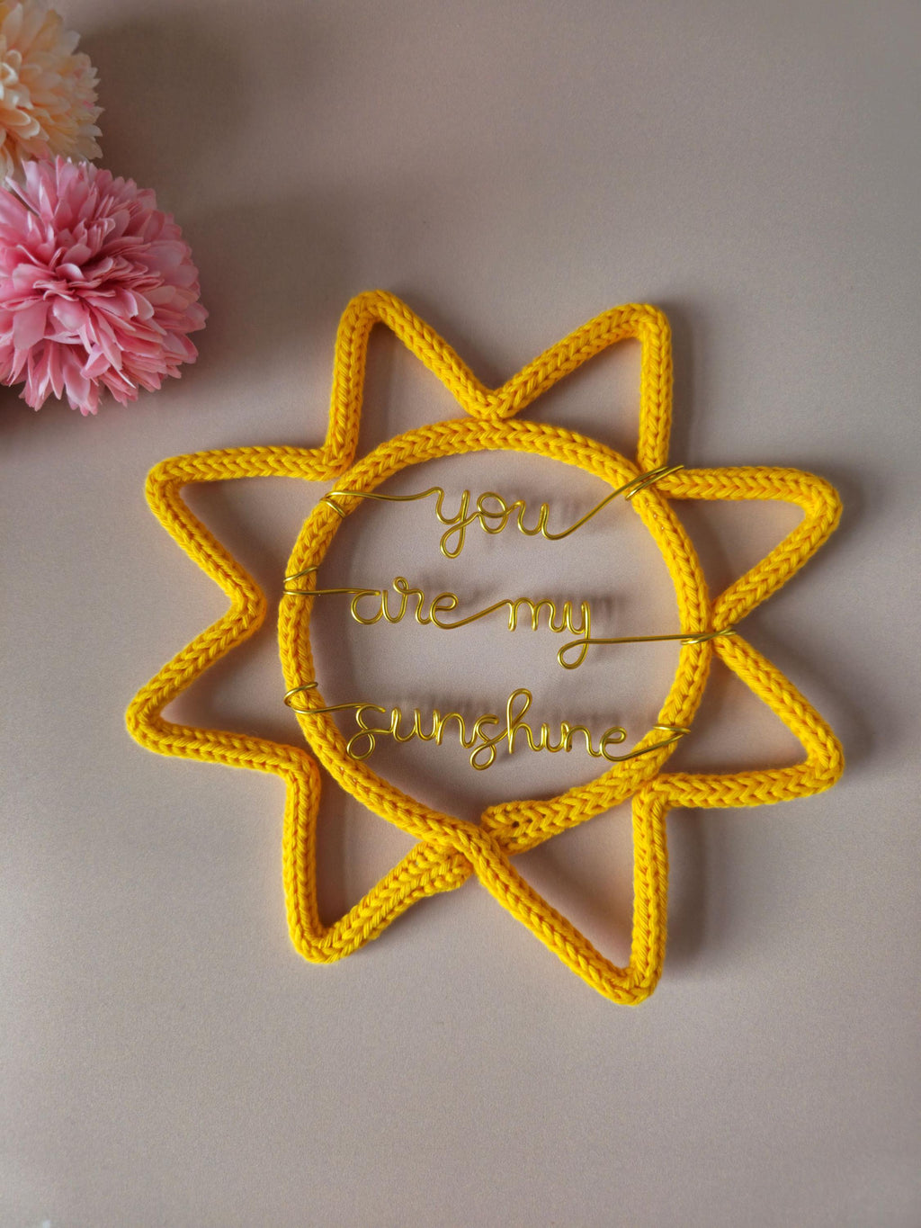 Yellow sun-shaped decorative item with 'you are my sunshine' text on a beige background with pink flowers.