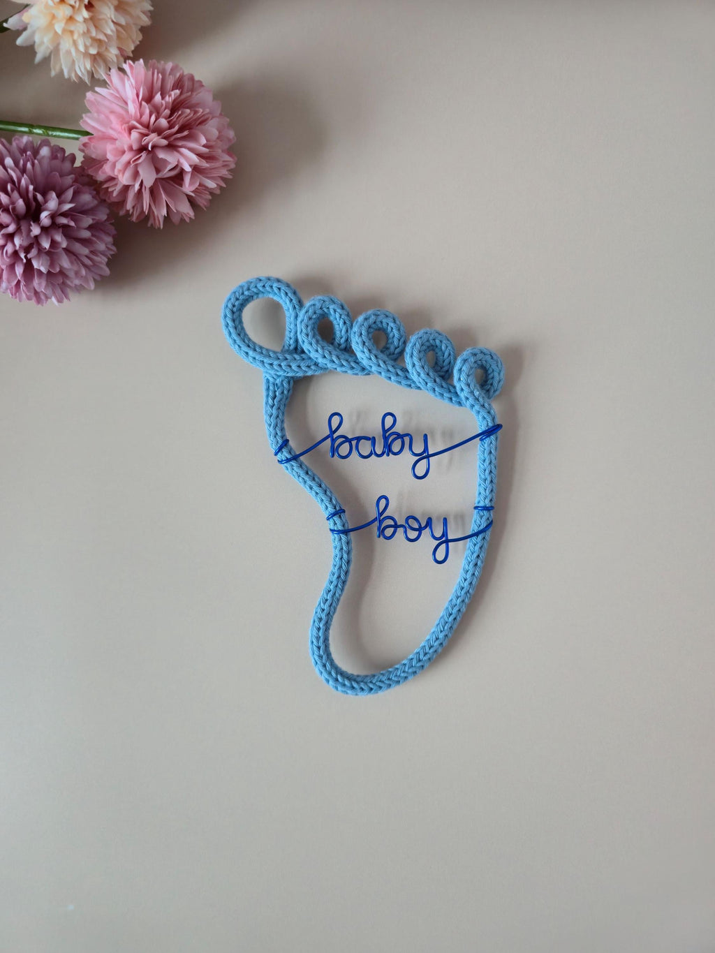 Blue knitted baby bootie with 'baby boy' text on a light background with flowers.
