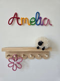 Rainbow knitted wire name sign reading Amelia displayed above a nursery shelf with a pink handmade flower detail.