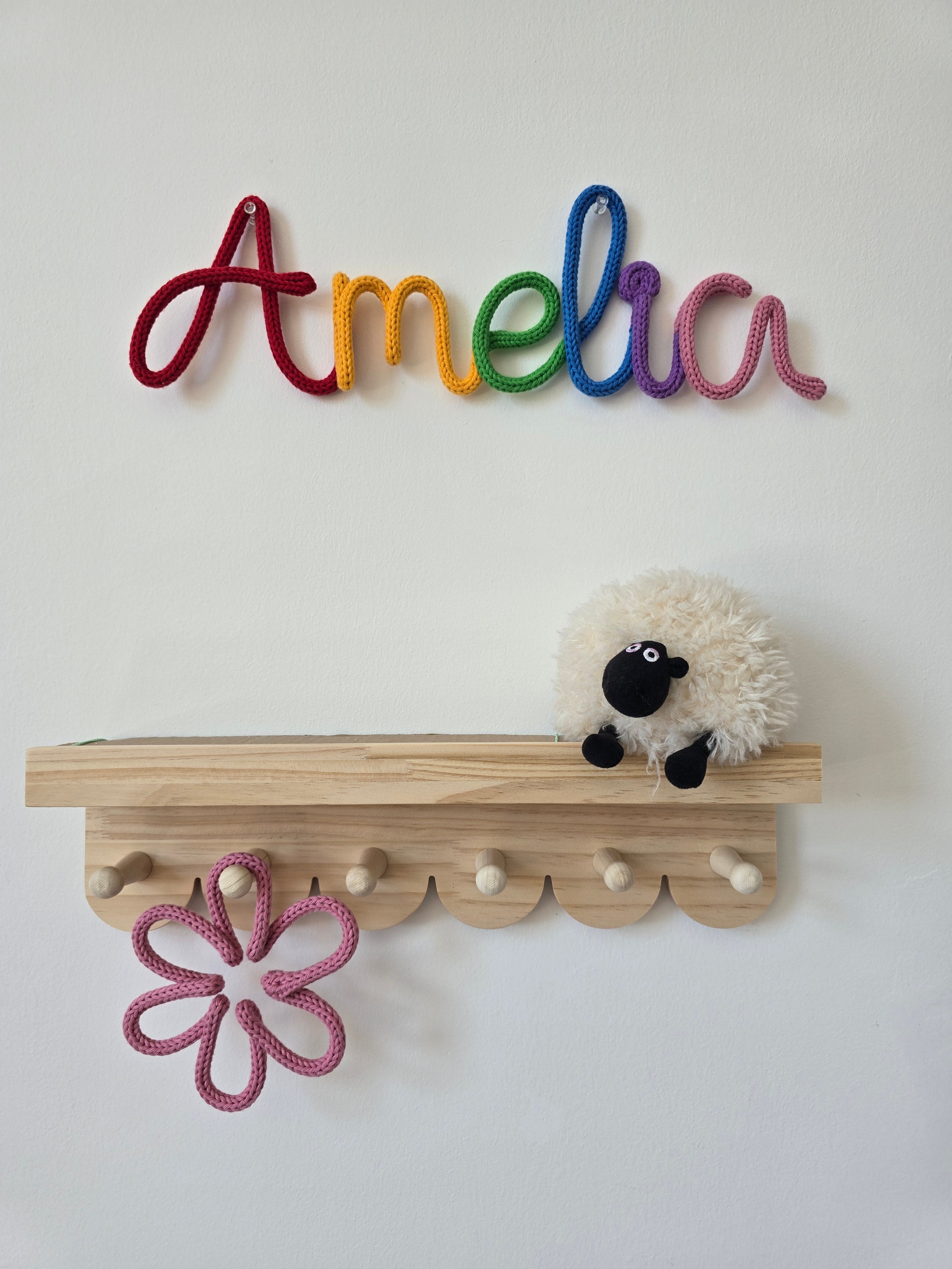 Rainbow knitted wire name sign reading Amelia displayed above a nursery shelf with a pink handmade flower detail.