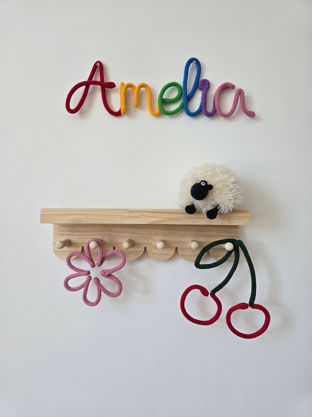 Rainbow personalised knitted wire name sign reading Amelia styled above a wooden shelf with handmade flower and cherry details for a child’s room.