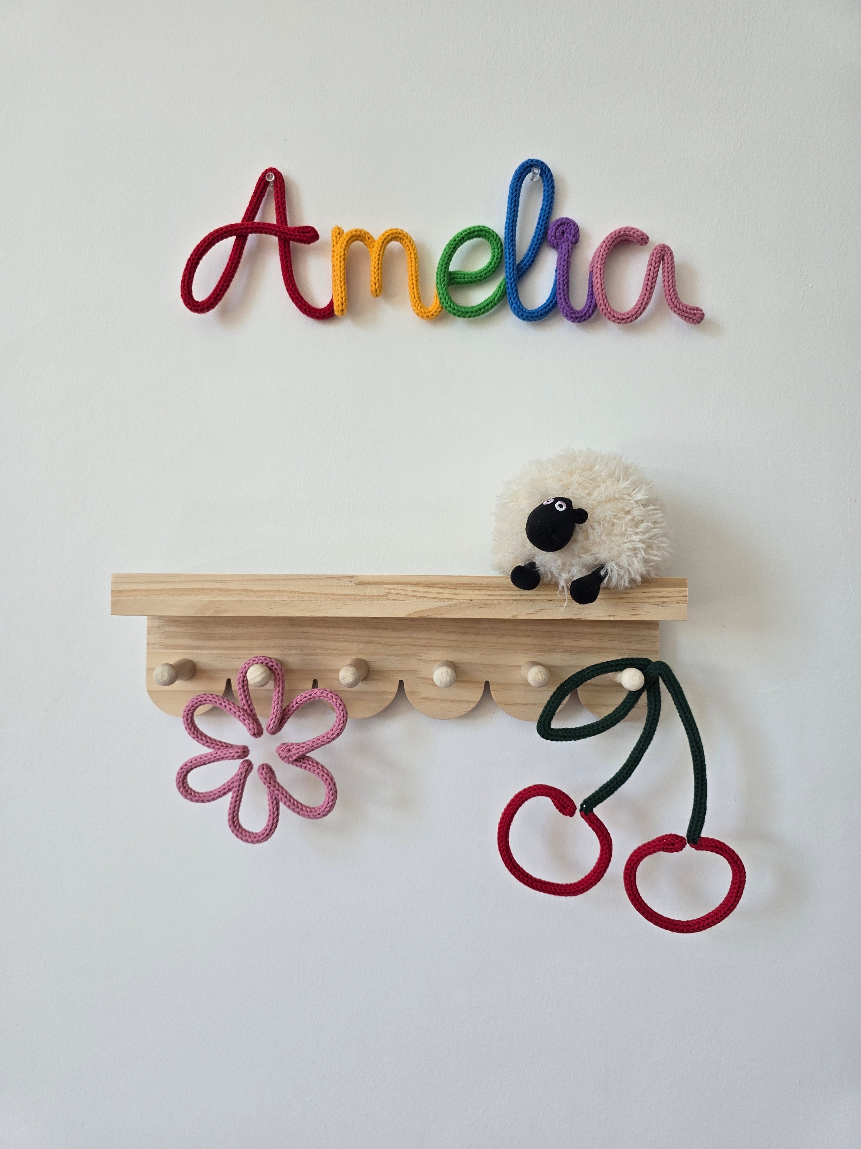 Rainbow personalised knitted wire name sign reading Amelia styled above a wooden shelf with handmade flower and cherry details for a child’s room.