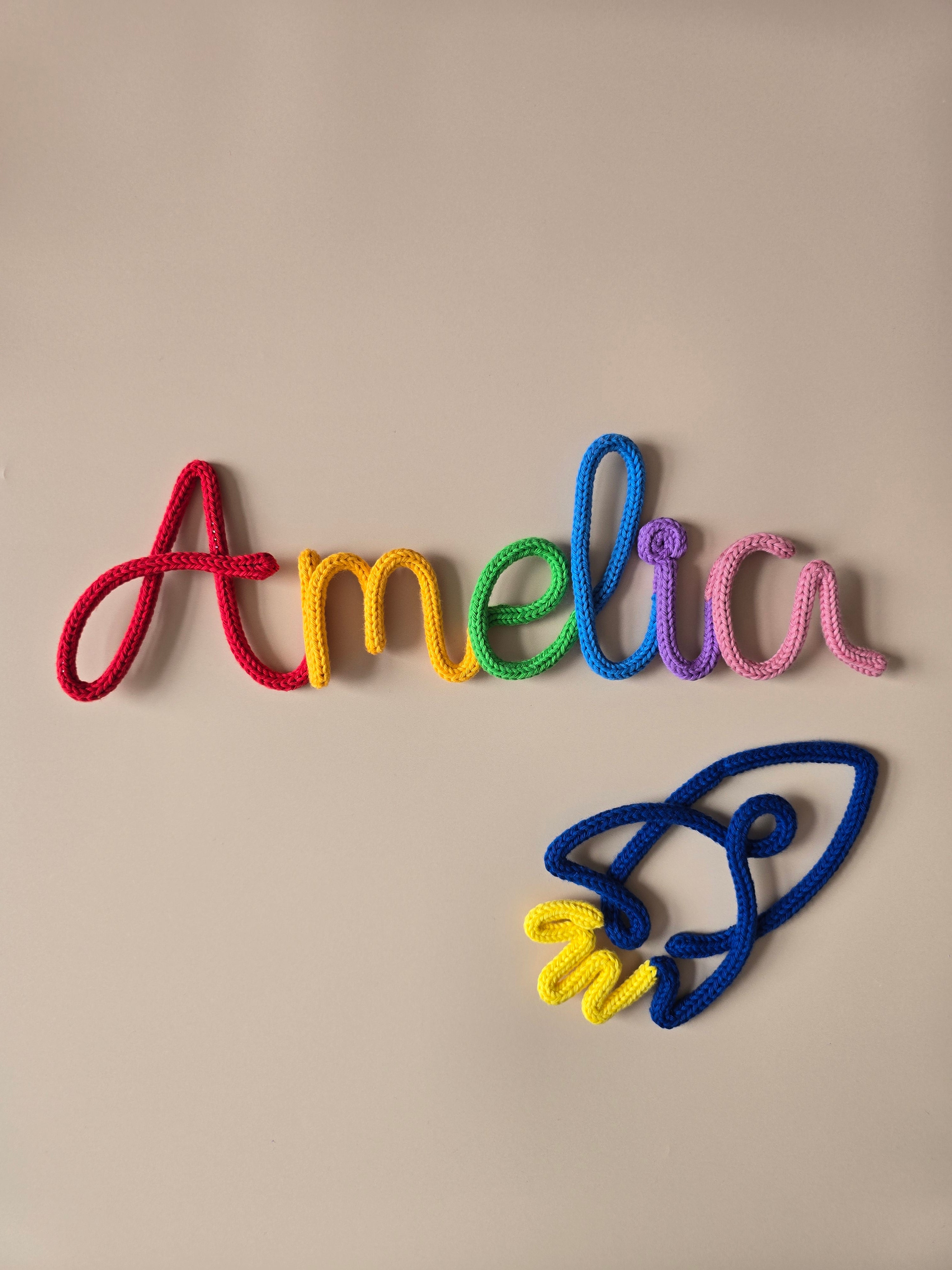 Rainbow personalised knitted wire name sign reading Amelia styled with a blue rocket wall decor shape.