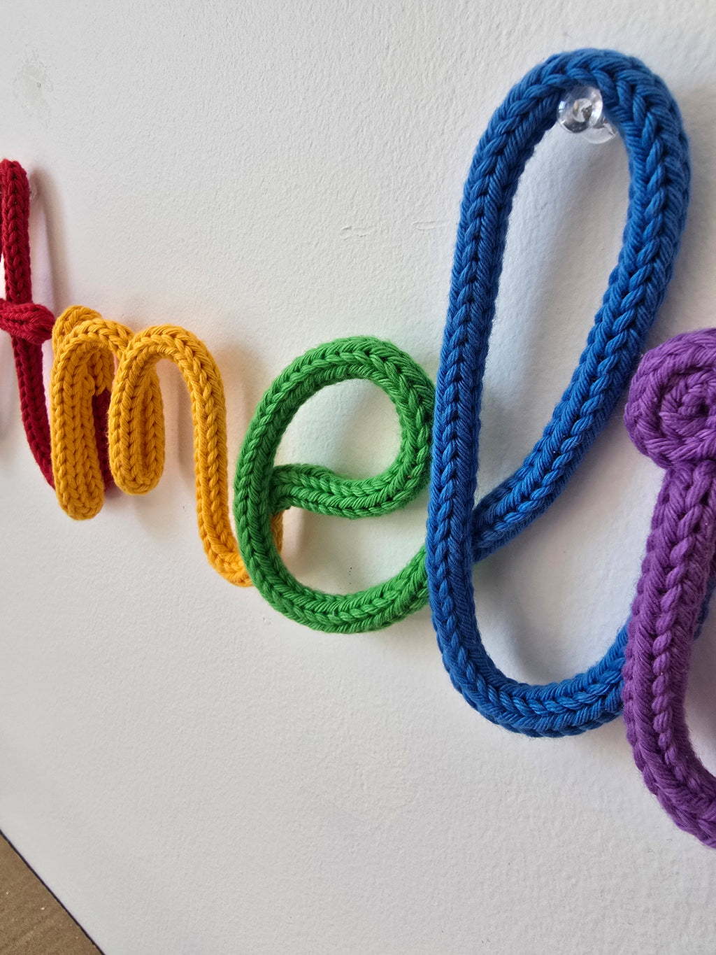Close-up of a rainbow knitted wire name sign showing soft cotton yarn texture and wall pin detail.