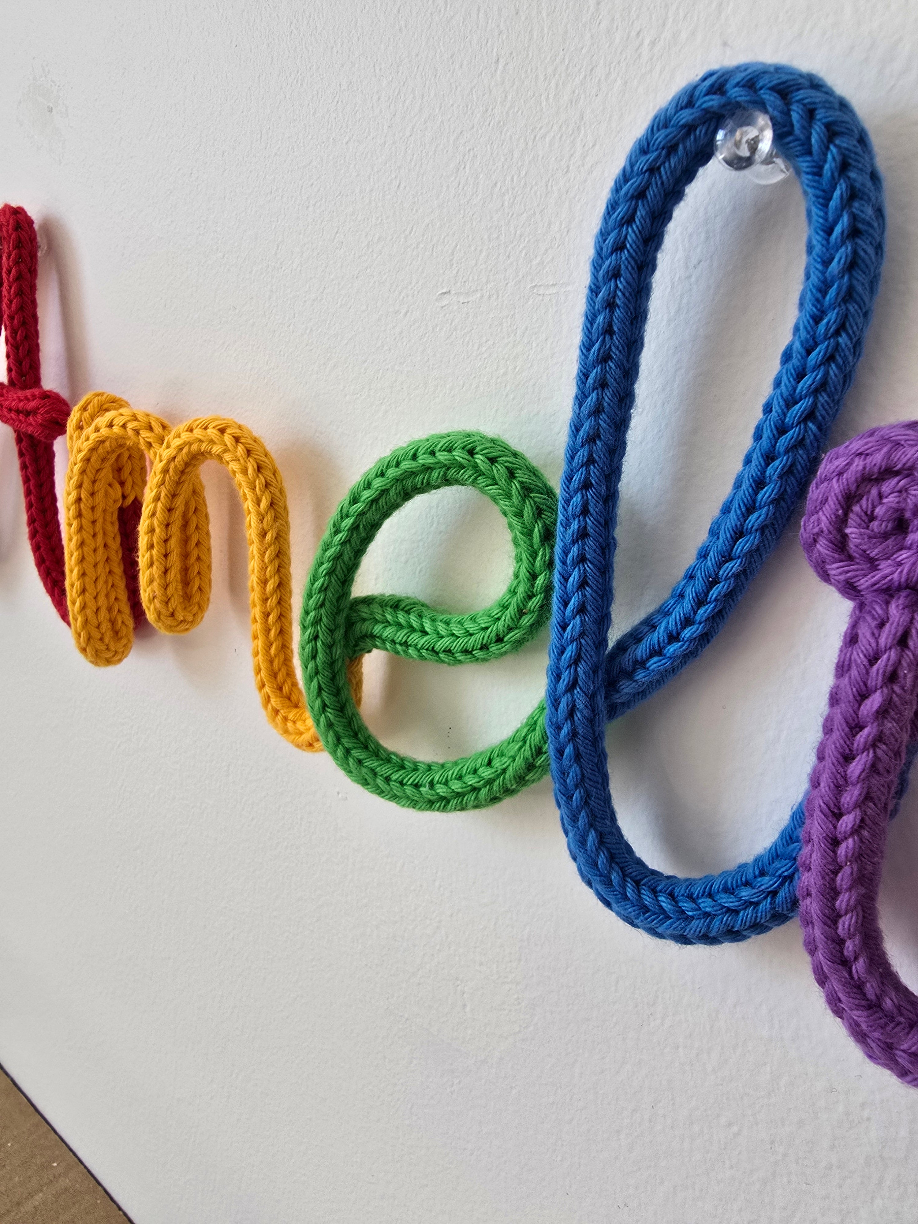 Close-up of a rainbow knitted wire name sign showing soft cotton yarn texture and wall pin detail.