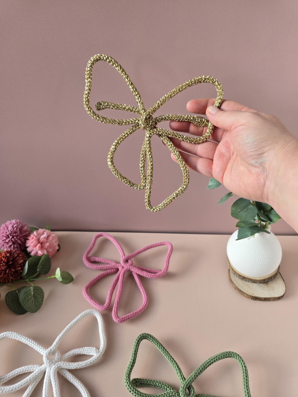Hand holding a gold butterfly made of rope on a pink background with other butterfly ropes in different colors.