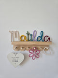 Multicoloured personalised knitted wire name sign reading Matilda, styled on a wooden shelf with matching flower, heart and cloud details for a child’s room.