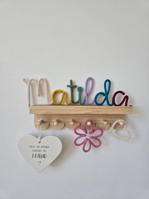 Multicoloured personalised knitted wire name sign reading Matilda, styled on a wooden shelf with matching flower, heart and cloud details for a child’s room.