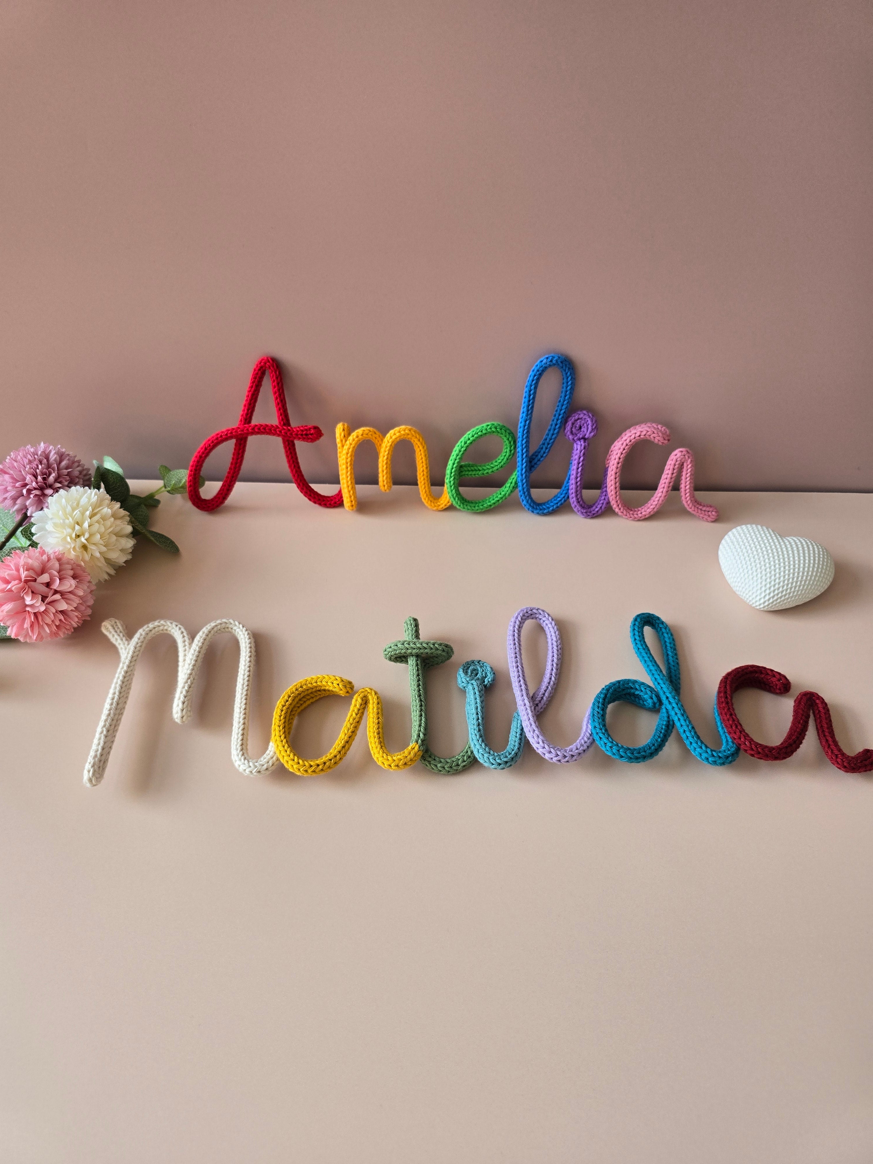 Two multicoloured personalised knitted wire name signs reading Amelia and Matilda, styled on a soft neutral backdrop with small handmade room details.