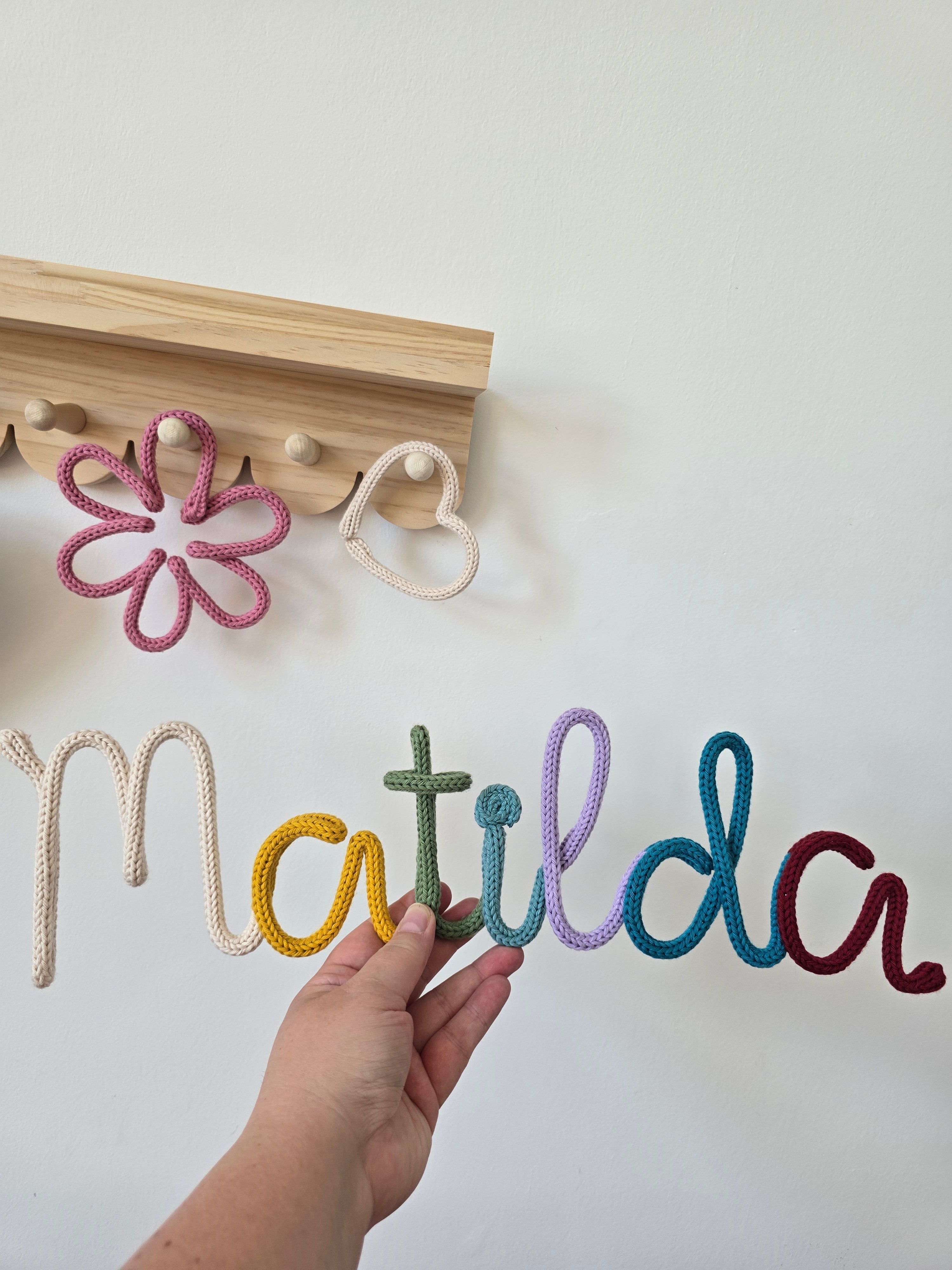 Personalised knitted wire name sign reading Matilda in the CraftyBabs signature multicoloured yarn mix, held by hand with a matching flower and heart detail.