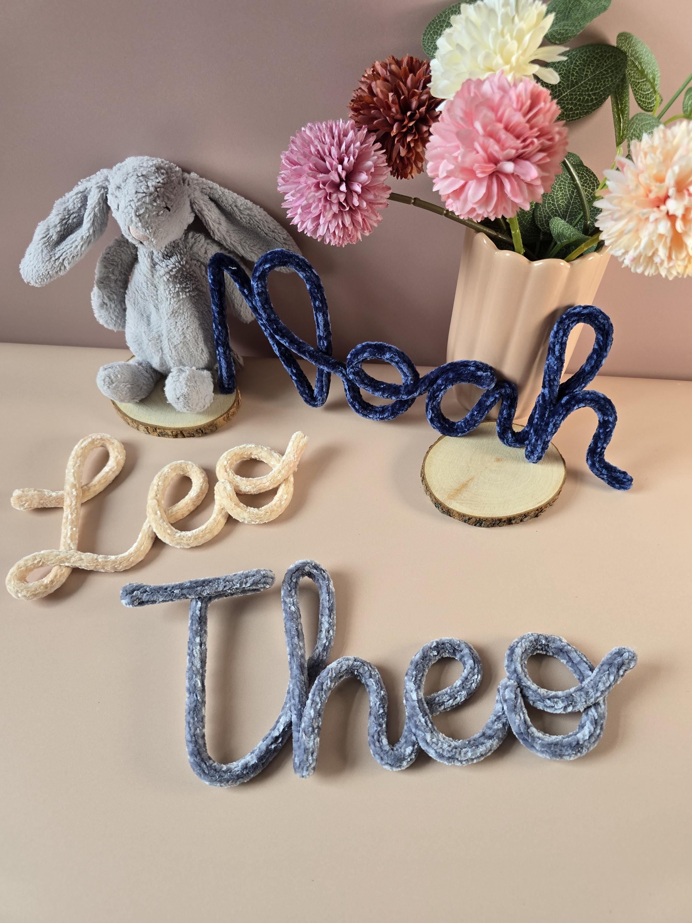 Decorative name signs with 'Noah', 'Lee', and 'Theo' next to a plush rabbit and flowers on a beige surface.