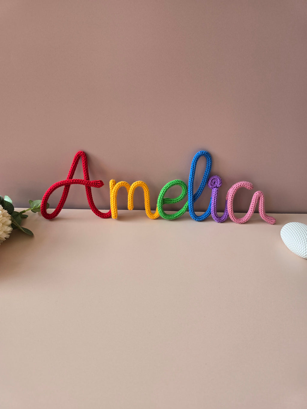 Rainbow knitted wire name sign reading Amelia in red, yellow, green, blue, purple and pink cotton yarn.