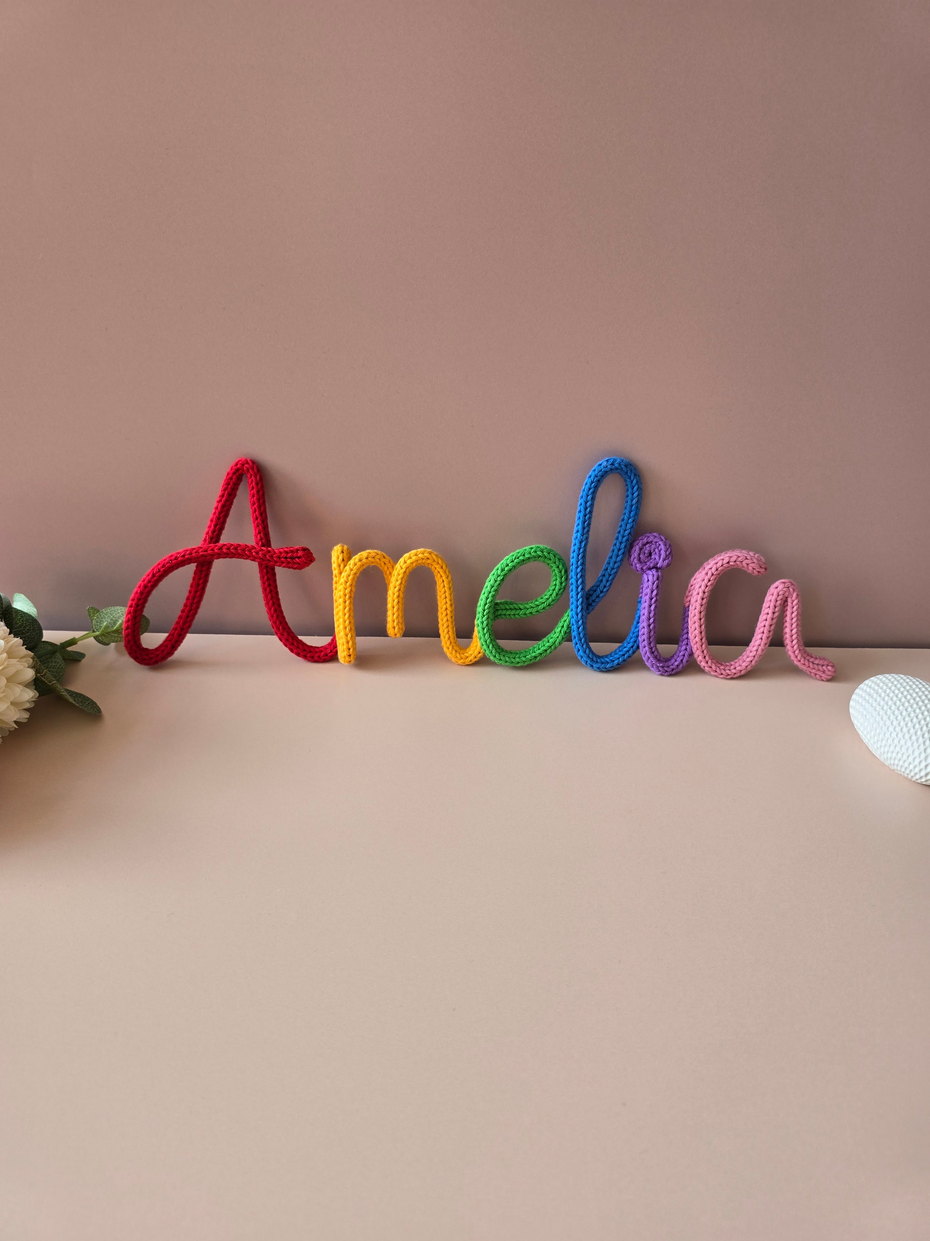 Rainbow knitted wire name sign reading Amelia in red, yellow, green, blue, purple and pink cotton yarn.