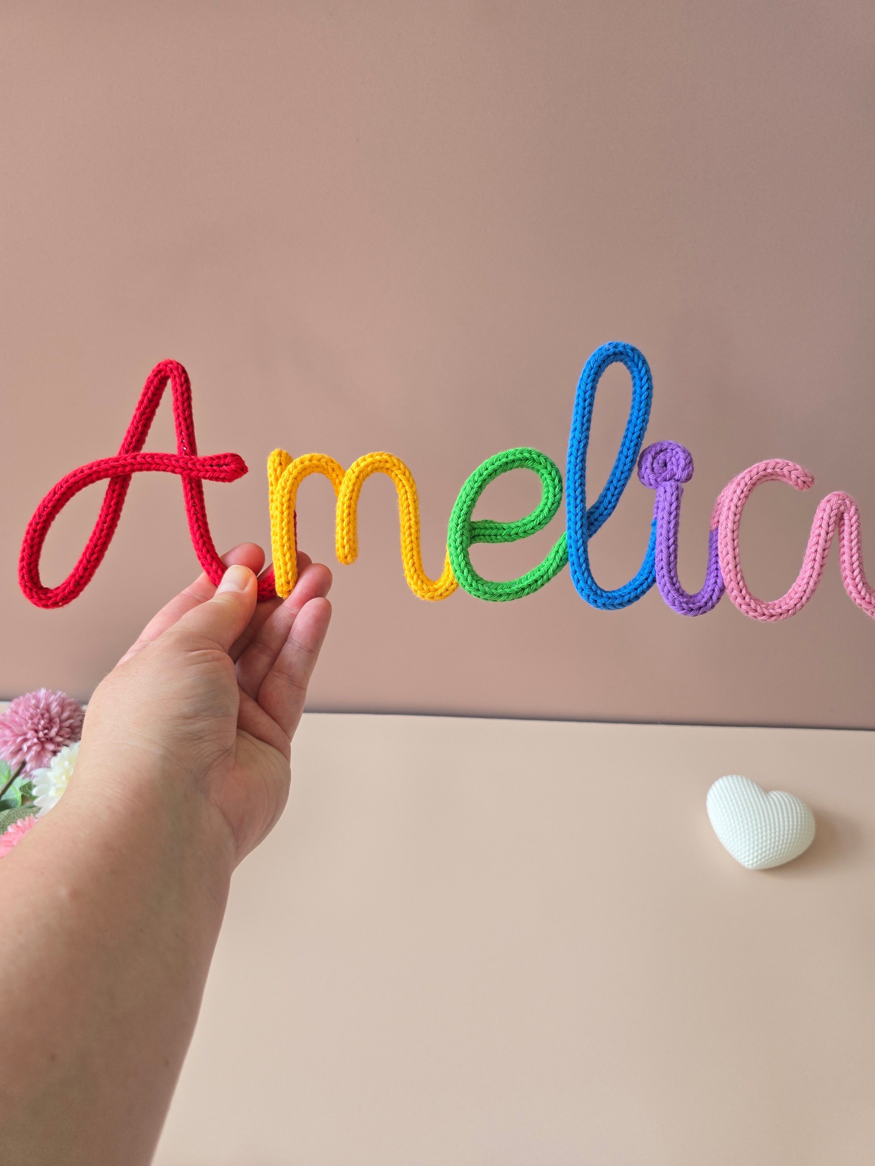 Hand holding a rainbow personalised knitted wire name sign reading Amelia, showing the handmade cotton yarn texture and scale.