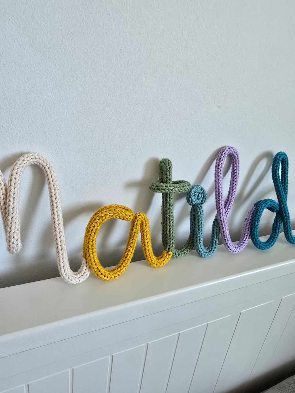 Close-up of a multicoloured knitted wire name sign showing the CraftyBabs signature colour mix in soft cotton yarn.