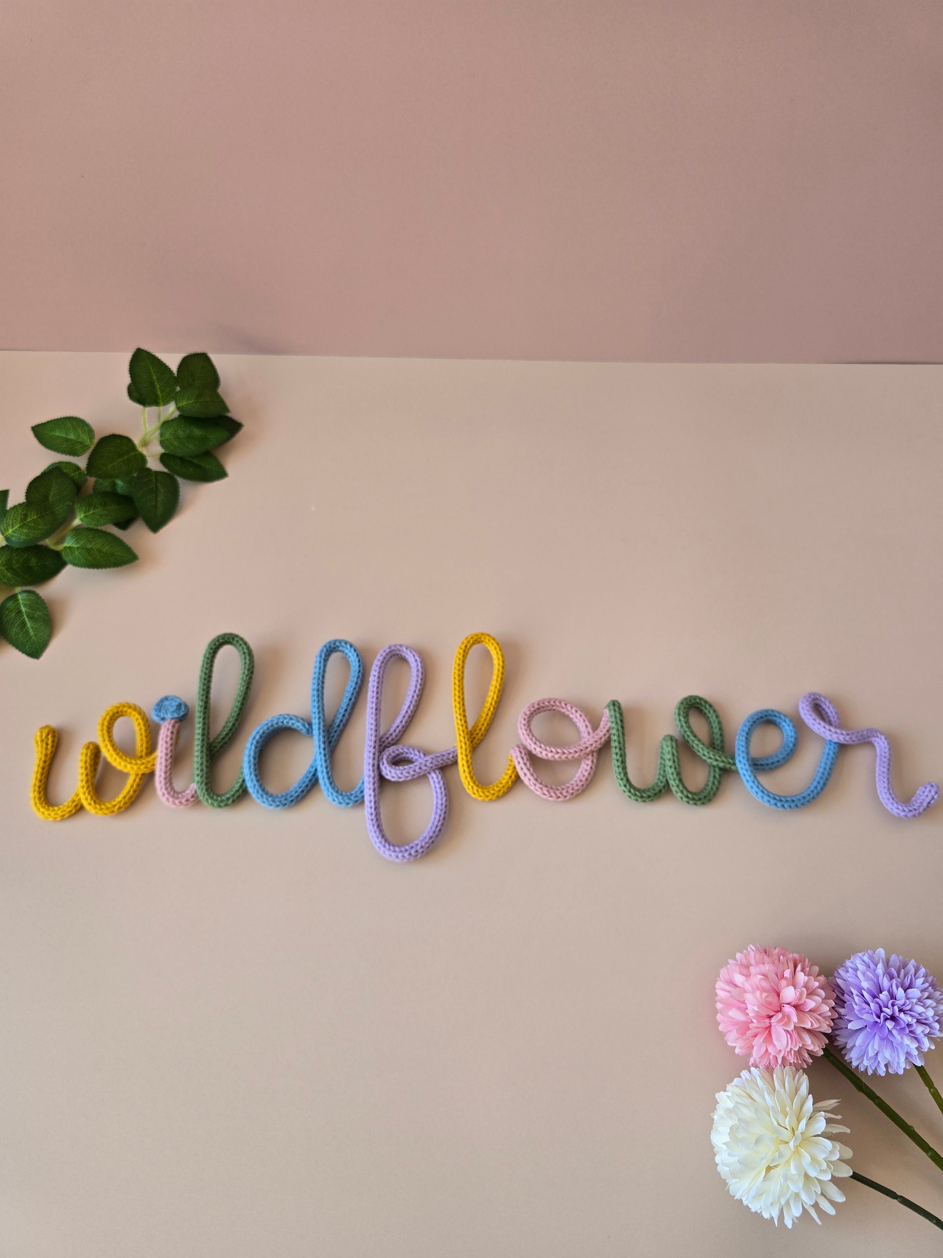 Multicolour Wildflower knitted-wire word styled with greenery and soft pom-pom flowers on a pink background.