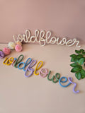 Cream and multicolour Wildflower knitted-wire wall words displayed together with flowers and greenery.